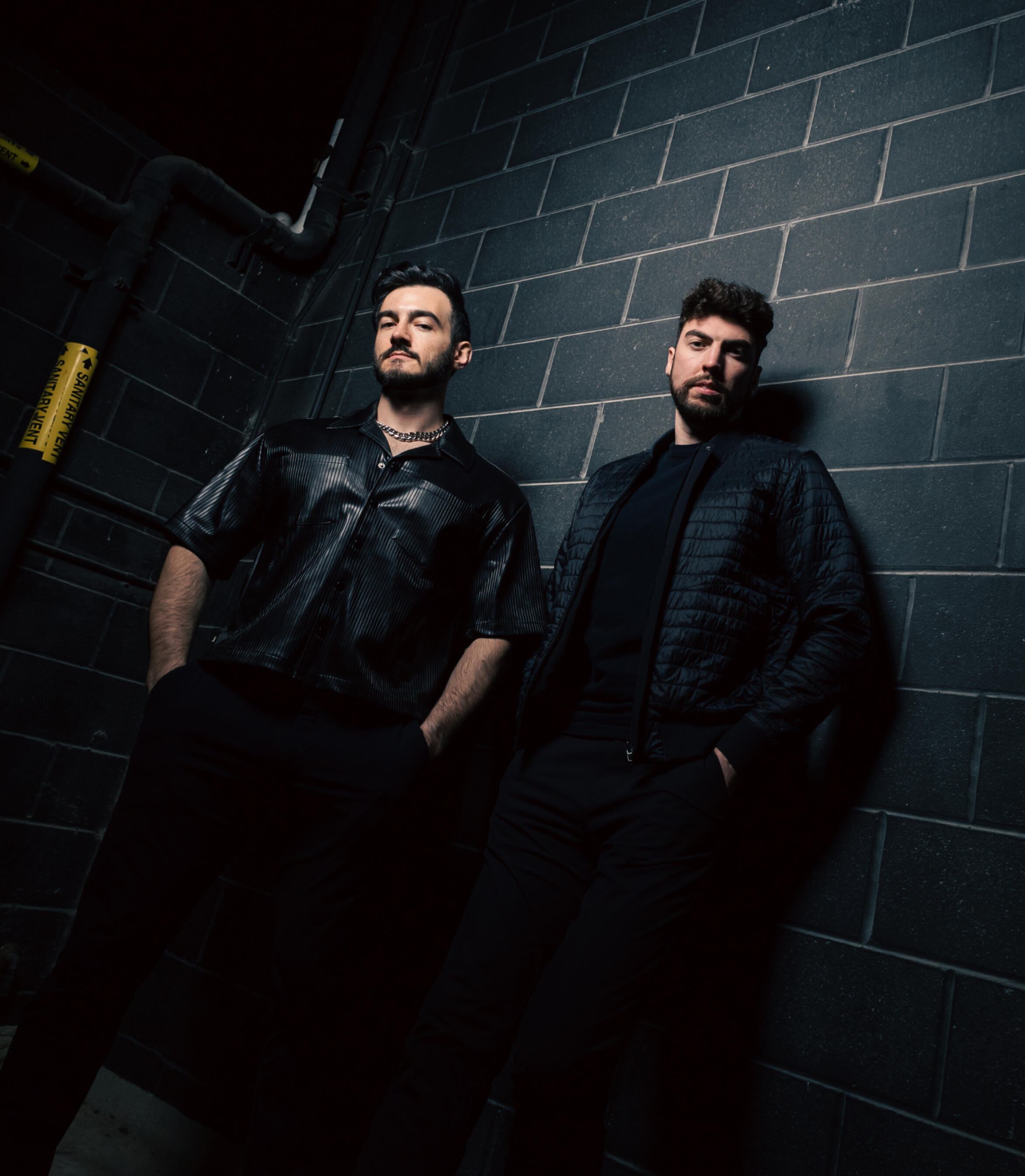Saint Stratus, a DJ duo of two men, wearing all black and standing near a black brick wall